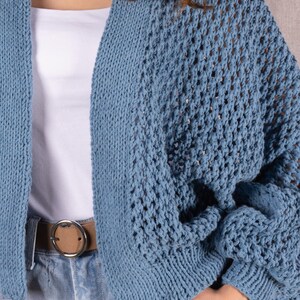 May include: A light blue knitted cardigan with an open weave design. The cardigan is worn over a white top and paired with blue jeans and a brown belt. The cardigan's sleeves are long and the overall style is casual.