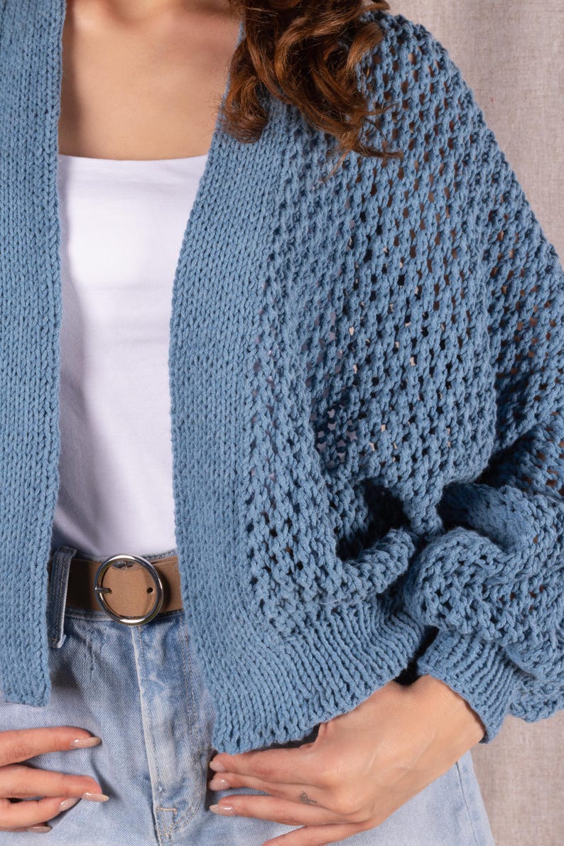 May include: A light blue knitted cardigan with an open weave design. The cardigan is worn over a white top and paired with blue jeans and a brown belt. The cardigan's sleeves are long and the overall style is casual.