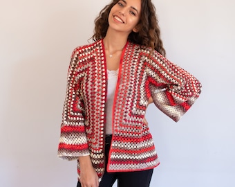 May include: A long-sleeved open-front cardigan made of crocheted granny squares in a repeating pattern of red, brown, white, and grey. The garment features a straight hemline, wide sleeves, and a solid red border trim along the front opening.