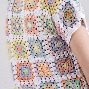 Puede incluir: Una blusa de manga corta confeccionada con m&uacute;ltiples cuadrados de ganchillo en tonos pastel de amarillo, rosa, azul y verde, unidos con hilo blanco. La prenda presenta un cuello redondo y un patr&oacute;n texturizado de calados.