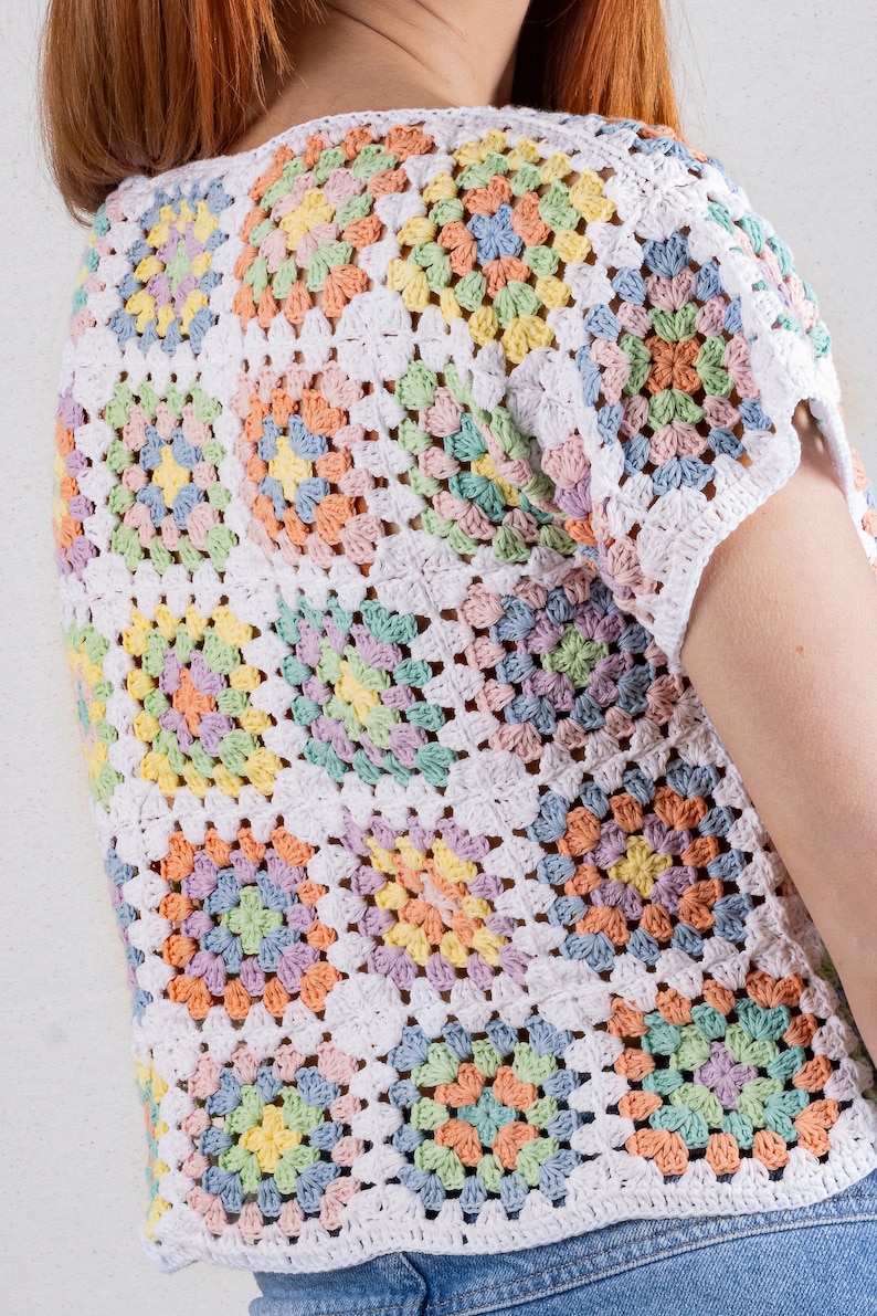 Puede incluir: Una blusa de manga corta confeccionada con m&uacute;ltiples cuadrados de ganchillo en tonos pastel de amarillo, rosa, azul y verde, unidos con hilo blanco. La prenda presenta un cuello redondo y un patr&oacute;n texturizado de calados.