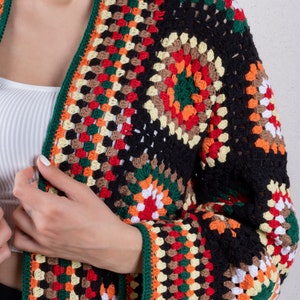 K&ouml;nnte beinhalten: Eine schwarze H&auml;keljacke mit bunten geometrischen Mustern in Rot, Orange, Gelb, Gr&uuml;n, Braun und Wei&szlig;. Die Jacke hat lange &Auml;rmel und einen tiefen V-Ausschnitt.