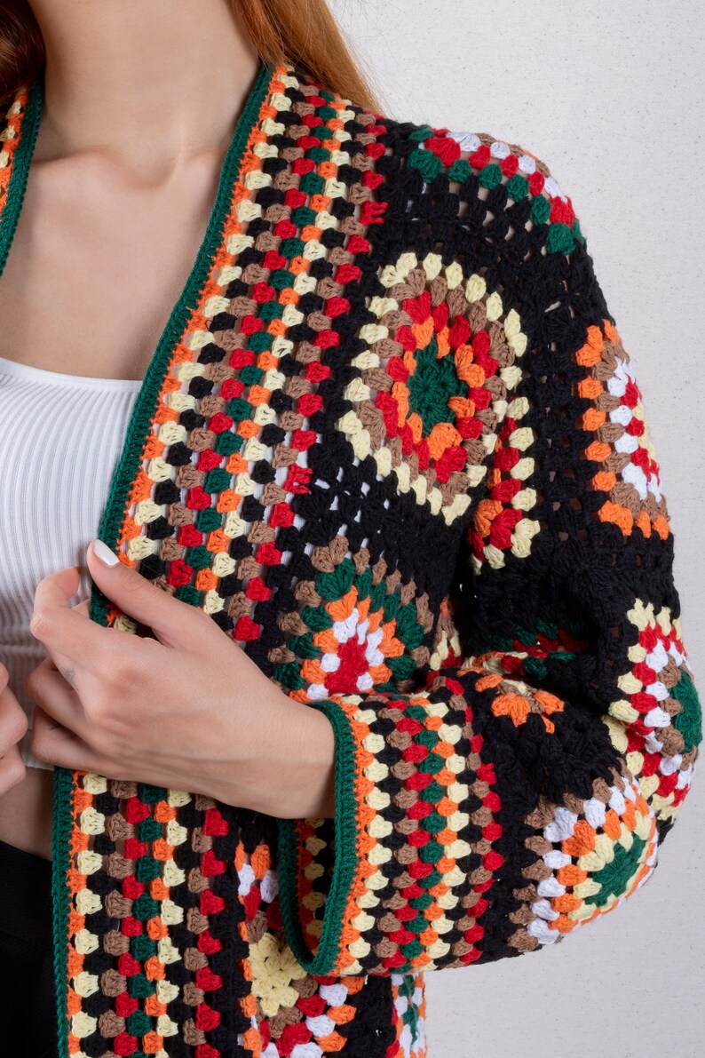 K&ouml;nnte beinhalten: Eine schwarze H&auml;keljacke mit bunten geometrischen Mustern in Rot, Orange, Gelb, Gr&uuml;n, Braun und Wei&szlig;. Die Jacke hat lange &Auml;rmel und einen tiefen V-Ausschnitt.