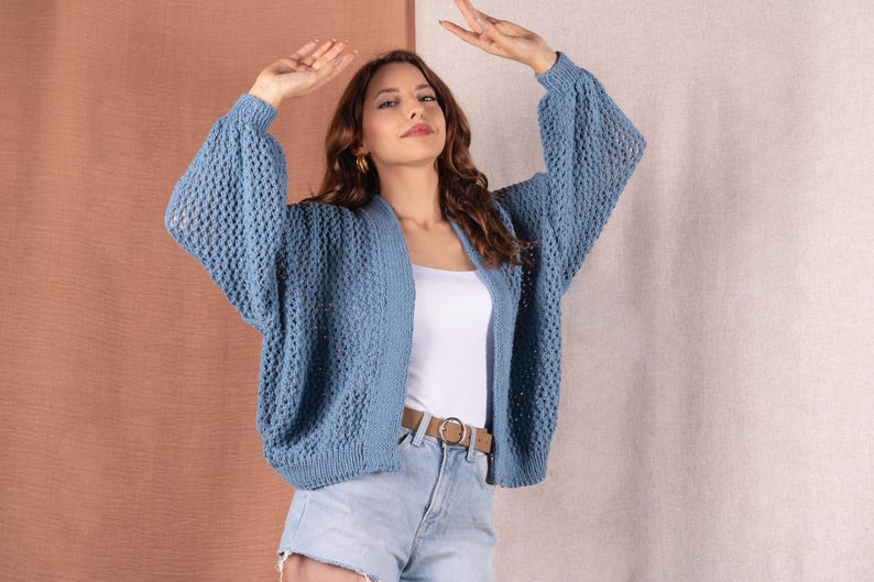 May include: A light blue open-front cardigan with a textured knit design. The cardigan has long, voluminous sleeves and is worn over a white top and light blue denim shorts. A brown belt with a gold buckle is visible.