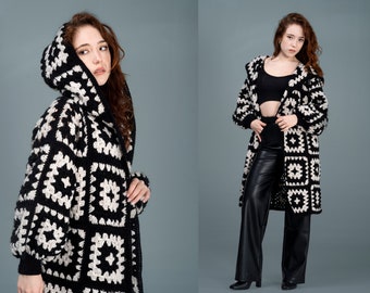 May include: A black and white crocheted cardigan with a hood. The cardigan is made of chunky yarn and has a square pattern. The model is wearing black pants.