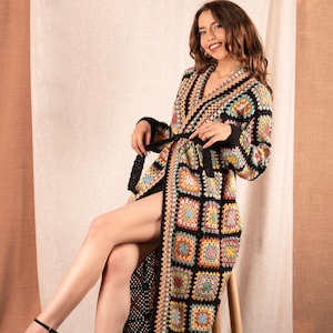 May include: A long, open-front crochet robe featuring a patchwork design of colorful squares. The robe has a black border and a matching belt. The model is sitting on a wooden chair.