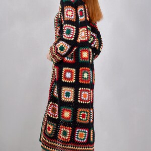 K&ouml;nnte beinhalten: Ein langer schwarzer H&auml;kelcardigan mit einem farbenfrohen geometrischen Muster. Der Cardigan hat eine lange, flie&szlig;ende Silhouette und ist aus verschiedenen Farben gefertigt, darunter Rot, Orange, Gelb, Gr&uuml;n und Wei&szlig;.
