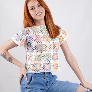 Puede incluir: Un top de ganchillo de manga corta construido a partir de una cuadr&iacute;cula de cuadrados de la abuela multicolores en tonos pastel de rosa, amarillo, azul y verde, bordeados con hilo blanco. La prenda presenta un cuello redondo y un dobladillo recto.