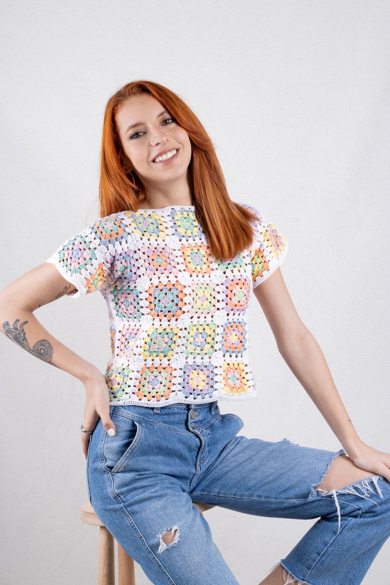 Puede incluir: Un top de ganchillo de manga corta construido a partir de una cuadr&iacute;cula de cuadrados de la abuela multicolores en tonos pastel de rosa, amarillo, azul y verde, bordeados con hilo blanco. La prenda presenta un cuello redondo y un dobladillo recto.