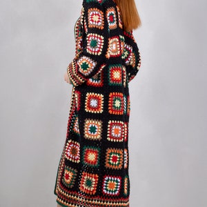 K&ouml;nnte beinhalten: Ein langer schwarzer H&auml;kelcardigan mit einem farbenfrohen geometrischen Muster. Der Cardigan hat eine lockere Passform und ist aus verschiedenen Farben gefertigt, darunter Rot, Orange, Gelb, Gr&uuml;n und Wei&szlig;.