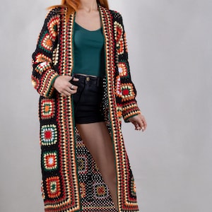 K&ouml;nnte beinhalten: Ein langer, schwarzer H&auml;kelcardigan mit bunten geometrischen Mustern. Der Cardigan hat einen tiefen V-Ausschnitt und ist vorne offen. Das Model tr&auml;gt den Cardigan &uuml;ber einem gr&uuml;nen Tanktop und schwarzen Shorts.