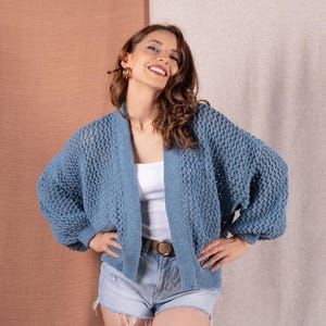 May include: A blue knit cardigan with a textured pattern. The open-front sweater has long, voluminous sleeves. The model is wearing a white top and denim shorts. The cardigan is a medium blue color.