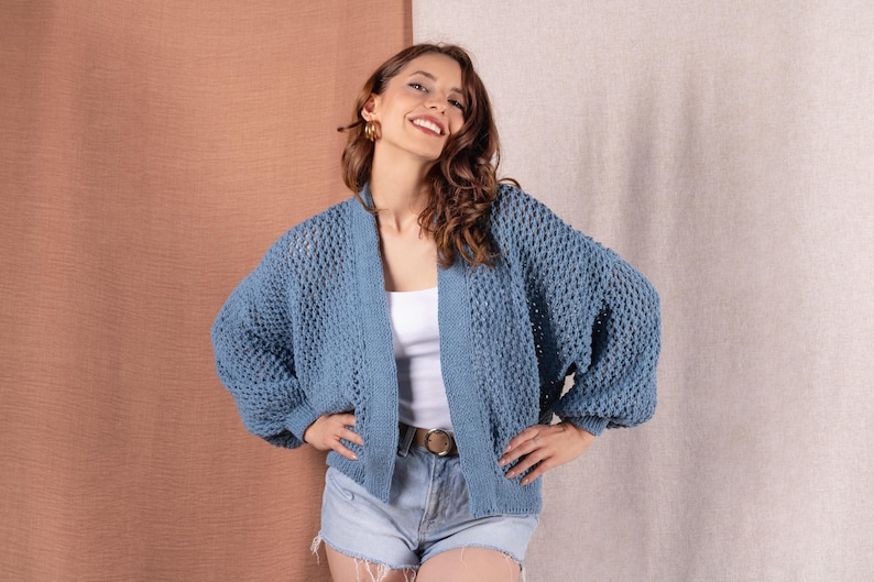 May include: A blue knit cardigan with a textured pattern. The open-front sweater has long, voluminous sleeves. The model is wearing a white top and denim shorts. The cardigan is a medium blue color.