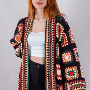 K&ouml;nnte beinhalten: Ein langer, schwarzer H&auml;kelcardigan mit bunten geometrischen Mustern. Der Cardigan hat einen weiten, offenen Vorderteil und lange &Auml;rmel.