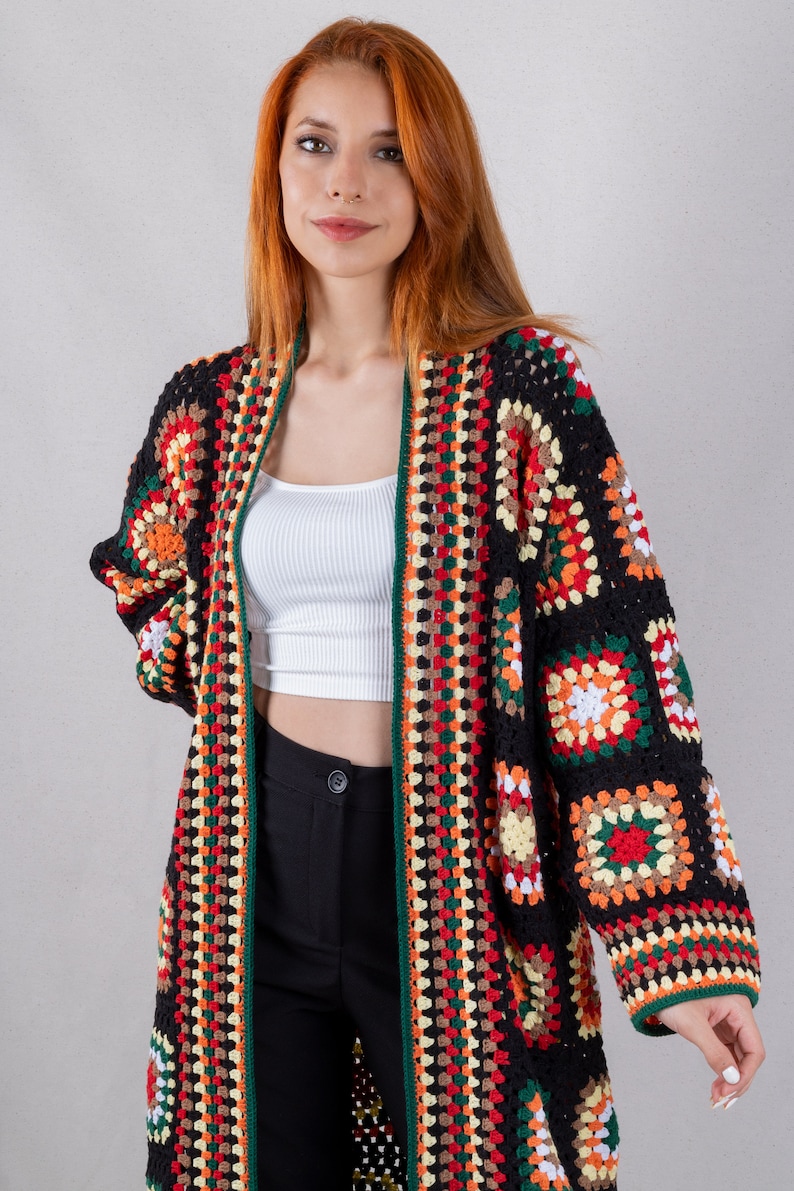 K&ouml;nnte beinhalten: Ein langer, schwarzer H&auml;kelcardigan mit bunten geometrischen Mustern. Der Cardigan hat einen weiten, offenen Vorderteil und lange &Auml;rmel.