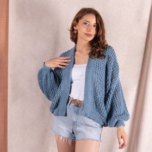 May include: A blue knit cardigan with a textured pattern and wide sleeves. The cardigan is worn over a white top and denim shorts. A brown belt with a gold buckle is visible.