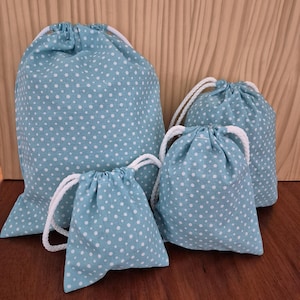 May include: Four drawstring bags in different sizes, arranged on a wooden surface. The bags are light blue with a white polka dot pattern. Each bag has a white drawstring closure.
