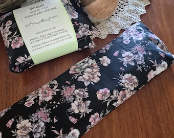 May include: A rectangular wheat heat or chill aches and pain relief bag featuring a black fabric with a floral pattern of pink and white flowers. A light green paper label is wrapped around the bag with instructions for microwave heating and freezer cooling.