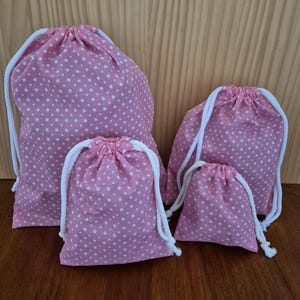 May include: A set of four pink drawstring bags in different sizes, patterned with white polka dots. Each bag features a white rope drawstring. They are displayed on a wooden surface, offering a coordinated storage solution.