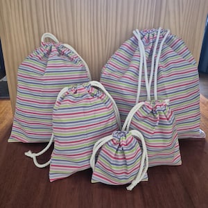 May include: Five drawstring bags of varying sizes, patterned with pink, green, blue, and purple horizontal stripes. Each bag has a white drawstring closure. The bags are designed for storage.
