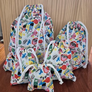 May include: A set of four drawstring bags in varying sizes, featuring a white fabric base with a colourful Play-Doh themed pattern. The print includes abstract shapes, dots, and the text "create" and "Play-Doh" in various colours and fonts.