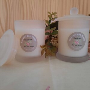 May include: Two white frosted glass jars with lids containing soy wax candles. The labels on the jars read "Coconut and Vanilla Handmade Soy Wax Candle".