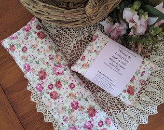 May include: Two floral patterned wheat and lavender heat or chill bags. The bags are filled with wheat and lavender and are designed to provide relief from aches and pains. The bags are on a wooden table with a wicker basket and a bouquet of pink flowers.