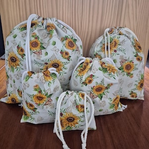 May include: A set of drawstring bags in different sizes, patterned with sunflowers, butterflies, and leaves on a light background. Each bag has a white drawstring. The bags are made of fabric and are displayed on a wooden surface.