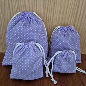 May include: Four drawstring bags in different sizes. The bags are light purple with a white polka dot pattern. Each bag has a white drawstring closure. The bags are displayed on a wooden surface.