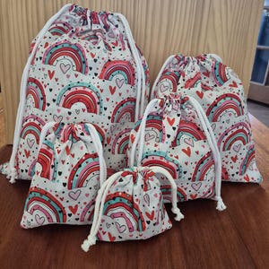May include: A set of four drawstring bags in varying sizes, each featuring a white fabric base printed with a repeating pattern of colourful rainbows in shades of red, pink, and teal, interspersed with small red and black hearts and white drawstrings.