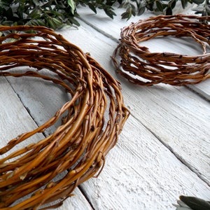May include: Two brown willow wreath forms on a white wooden surface. The wreath forms are made of woven willow branches.