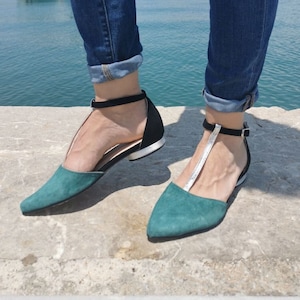 May include: Teal and black flats with a silver strap and ankle strap. The shoes have a pointed toe and a black heel. The flats are worn with blue jeans. The background shows a body of water and a building.