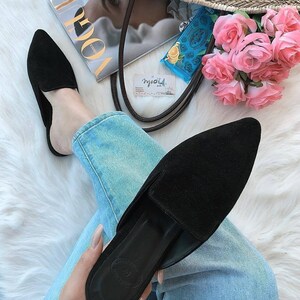 Puede incluir: Un par de mules de punta de color negro de ante con un tac&oacute;n bajo. Los zapatos son usados por una persona que lleva pantalones vaqueros azules y est&aacute; sentada en una superficie blanca y esponjosa.