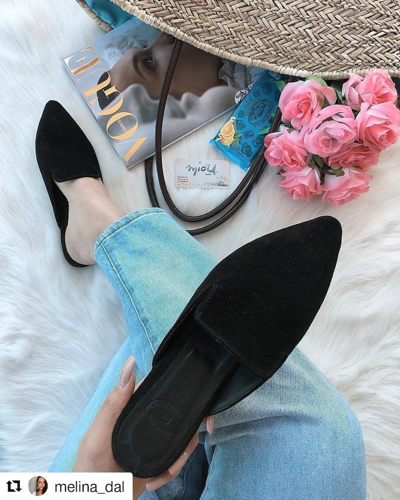 Puede incluir: Un par de mules de punta de color negro de ante con un tac&oacute;n bajo. Los zapatos son usados por una persona que lleva pantalones vaqueros azules y est&aacute; sentada en una superficie blanca y esponjosa.