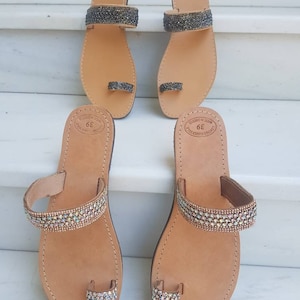 May include: Two pairs of leather sandals with jeweled straps. The sandals have a toe ring and are made in Greece. The sandals are brown with a light brown sole. One pair has iridescent jewels and the other pair has black jewels.