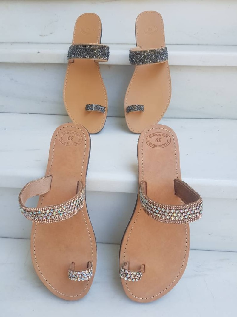 May include: Two pairs of leather sandals with jeweled straps. The sandals have a toe ring and are made in Greece. The sandals are brown with a light brown sole. One pair has iridescent jewels and the other pair has black jewels.