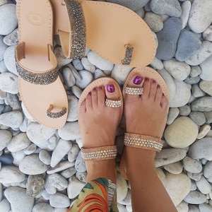 May include: A pair of tan leather sandals with jeweled straps. The sandals have a toe ring and are adorned with rhinestones. The sandals are on a bed of small, smooth stones.