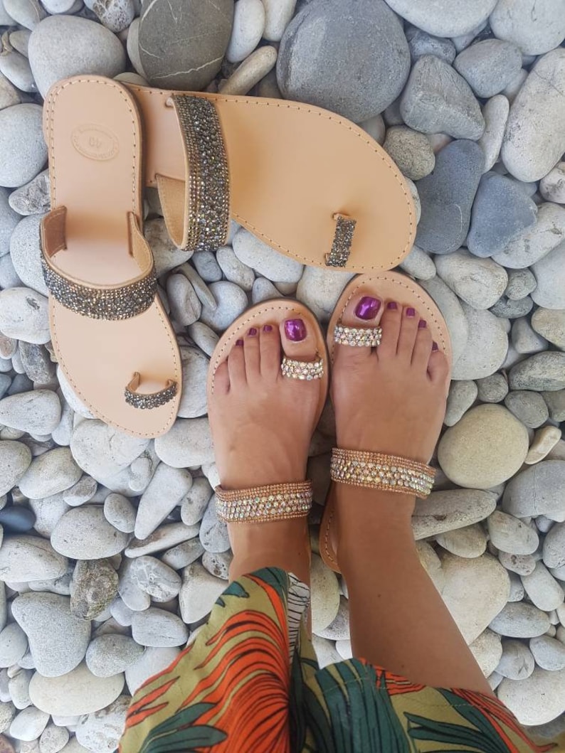 May include: A pair of tan leather sandals with jeweled straps. The sandals have a toe ring and are adorned with rhinestones. The sandals are on a bed of small, smooth stones.