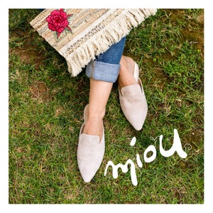 Puede incluir: Un par de mules de punta de color beige claro de gamuza con un tac&oacute;n bajo. Los zapatos son usados por una persona sentada en una superficie cubierta de hierba. La palabra "miou" est&aacute; escrita en cursiva blanca sobre la hierba.