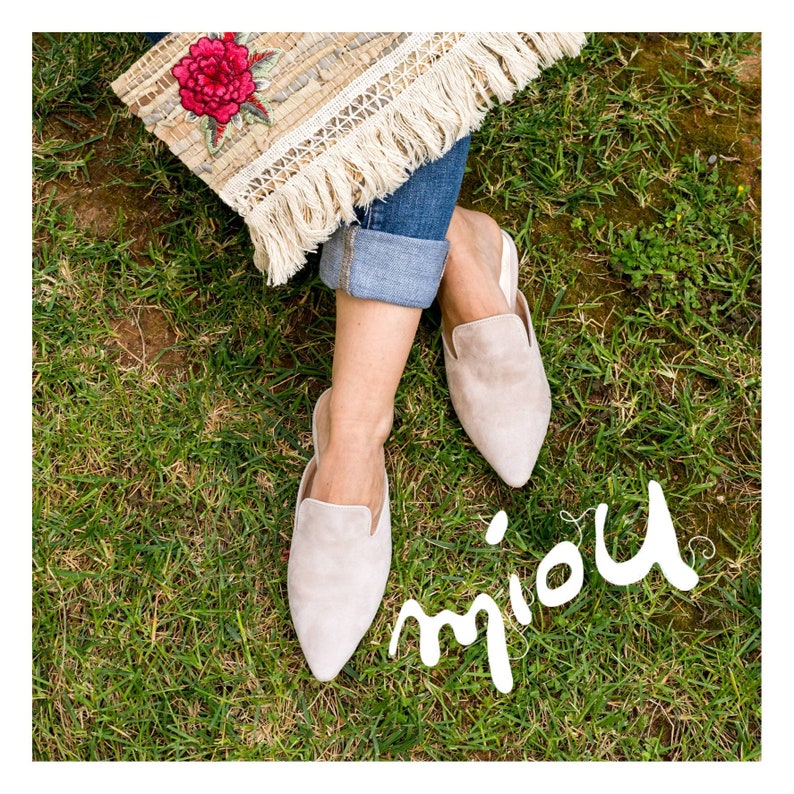 Puede incluir: Un par de mules de punta de color beige claro de gamuza con un tac&oacute;n bajo. Los zapatos son usados por una persona sentada en una superficie cubierta de hierba. La palabra "miou" est&aacute; escrita en cursiva blanca sobre la hierba.