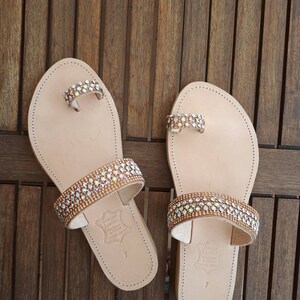 May include: A pair of beige leather sandals with two straps adorned with rows of iridescent rhinestones. The sandals have a toe ring and the text "Genuine Leather Handmade in Greece" is printed on the sole.