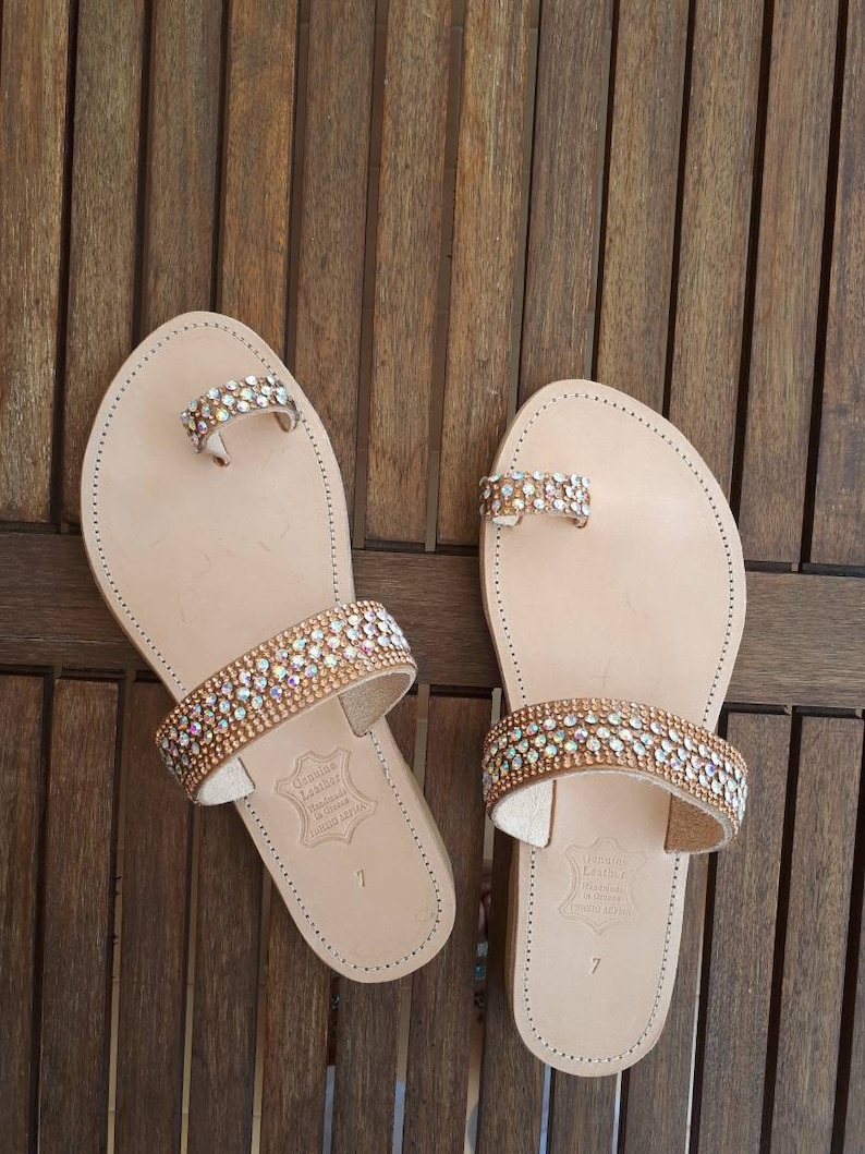 May include: A pair of beige leather sandals with two straps adorned with rows of iridescent rhinestones. The sandals have a toe ring and the text "Genuine Leather Handmade in Greece" is printed on the sole.