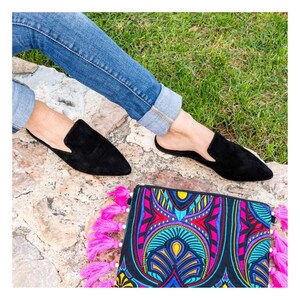 Puede incluir: Un par de mules de ante negro de punta puntiaguda con ribete de borlas. Los zapatos los lleva una persona que lleva pantalones vaqueros azules. Los zapatos est&aacute;n sobre una superficie de piedra con un bolso de mano estampado de colores con borlas rosas.