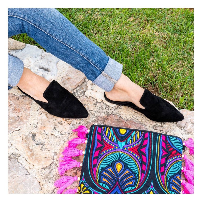 Puede incluir: Un par de mules de ante negro de punta puntiaguda con ribete de borlas. Los zapatos los lleva una persona que lleva pantalones vaqueros azules. Los zapatos est&aacute;n sobre una superficie de piedra con un bolso de mano estampado de colores con borlas rosas.