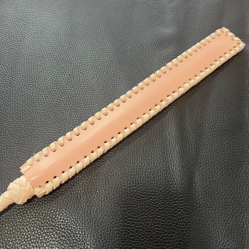May include: A long, light brown leather whip with a braided handle and decorative stitching along the edges.
