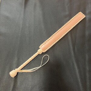 May include: A light pink leather paddle with a braided handle and a loop at the end. The paddle is on a black surface.