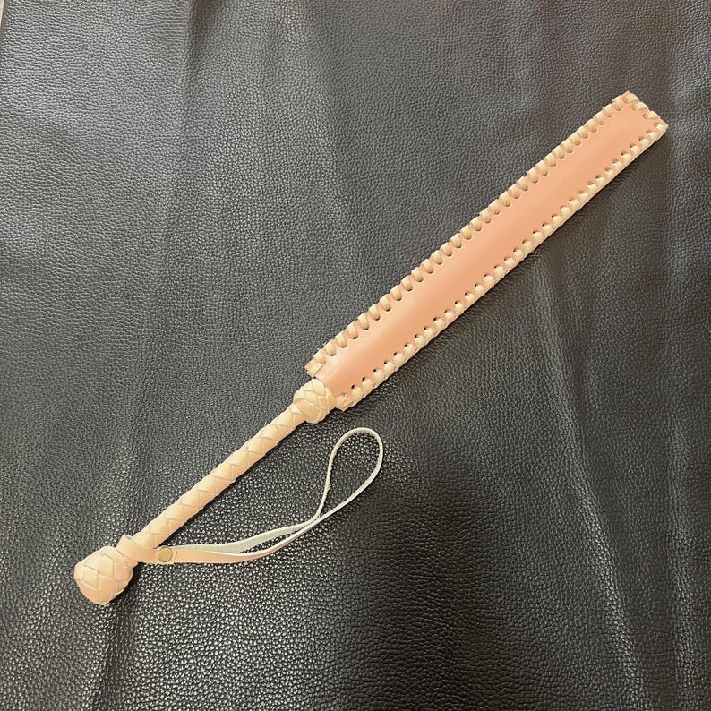 May include: A light pink leather paddle with a braided handle and a loop at the end. The paddle is on a black surface.