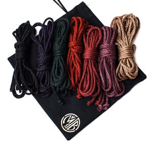 Pode incluir: Conjunto de seis cordas coloridas em uma bolsa de cordão preto. As cordas são em tons de preto, verde escuro, vermelho, bordô e marrom.