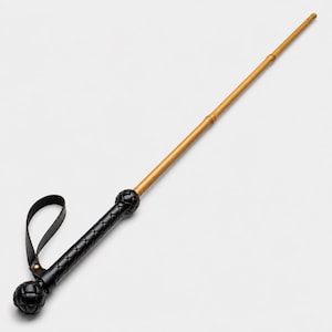 May include: A long, slender, golden-brown rod attached to a black handle. The handle features a woven leather texture, a spherical pommel at the base, and a black leather wrist strap secured with a small gold-toned metal rivet near the top.