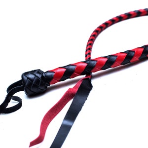 Op de afbeelding: Een zwarte en rode leren zweep met een gevlochten ontwerp. De zweep heeft een zwart leren handvat en een rood-zwarte gevlochten riem.